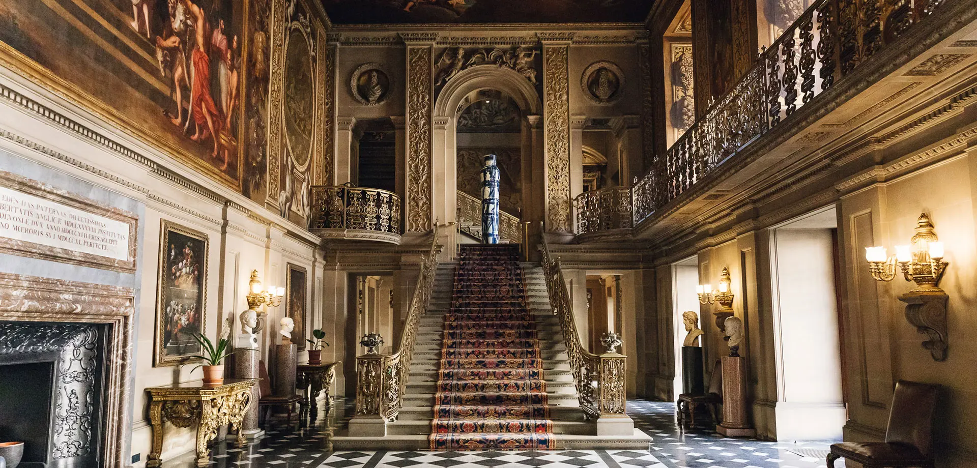 A grand hall with a black and white checkerboard floor, frescos on the walls and ceiling, and a central staircase leading up to a landing.